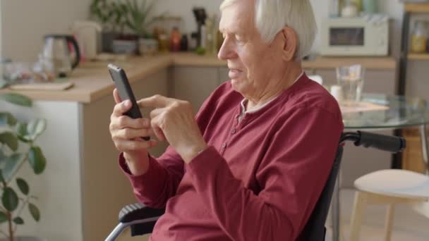 Naklonit se záběr seniora na invalidním vozíku pomocí smartphonu při rolování na sociálních sítích doma - Záběry, video