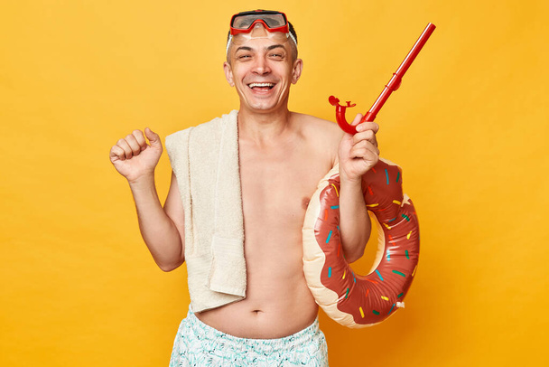 Happy cheerful man in diving goggles and snorkel holding donut rubber ring isolated on yellow background rejoicing his simmer activity clenched fists smiling. - Foto, Imagem