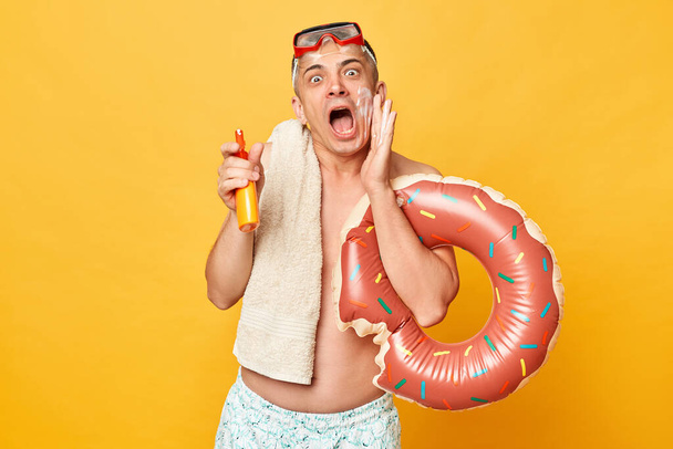 Excited man in diving goggles and snorkel holding donut rubber ring isolated on yellow background holding sunscreen making announcement screaming with hand near mouth. - Fotó, kép
