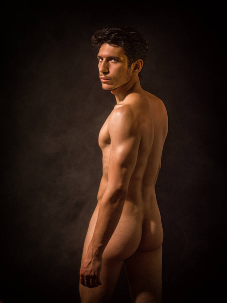 Portrait of naked handsome young man with languishing look covering crotch with his hands. In a studio with a dark backdrop, an athletic, handsome young man stands completely nude looking at camera. - Foto, afbeelding