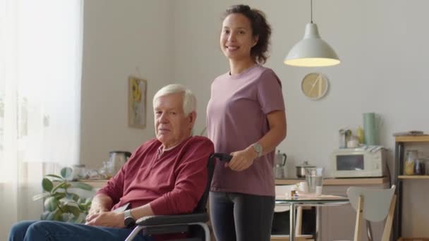 Medium long shot of cheerful female caregiver and elderly man in wheelchair posing together for camera during day at home - Footage, Video