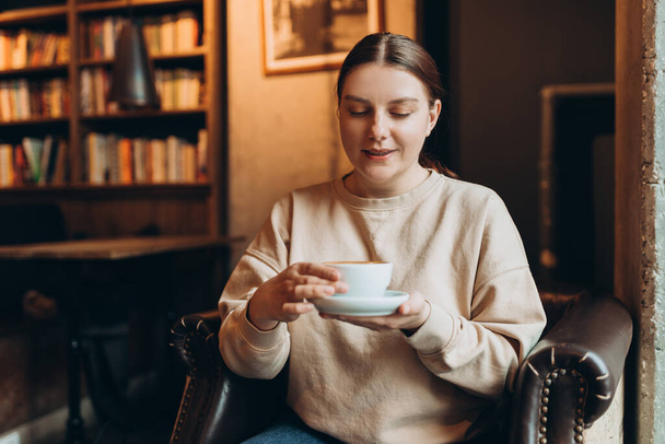 Portrét radostné mladé ženy, která si doma vychutnává šálek kávy. Usmívající se hezká dívka pije horký nápoj v kavárně v zimě. Nadšené ženy nosí svetr v podzimním dni. - Fotografie, Obrázek
