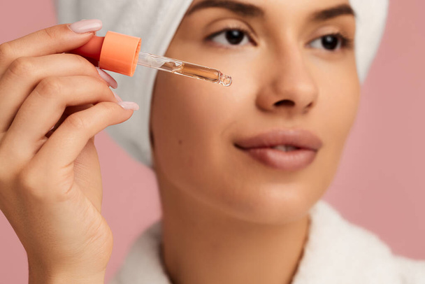 Soft focus of crop young female in bathrobe and towel on head applying rejuvenating serum with dropper during beauty procedure against pink background - Foto, Bild