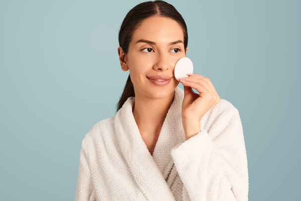 Smiling young brunette wearing white bathrobe putting round cotton pad on cheek while removing makeup against turquoise background - Foto, Bild