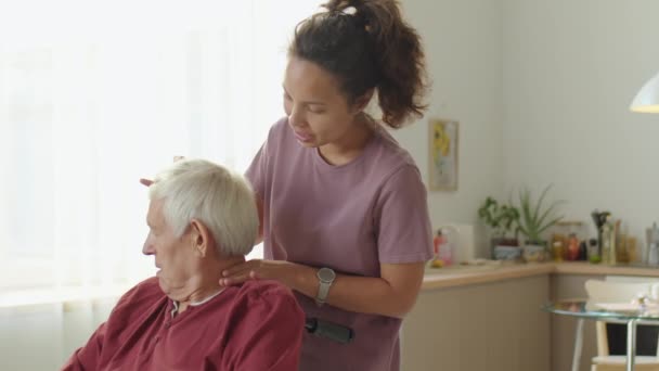 Vrouwelijke fysiotherapeut die het hoofd van een oudere man in een rolstoel draait en spreekt terwijl hij hem medische zorg thuis verleent - Video