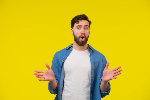 A portrait of a bearded young adult man with open palms sideways. Looking shocked and speechless. Taken by surprise with eyes and mouth wide opened. Isolated on yellow background. - Фото, изображение