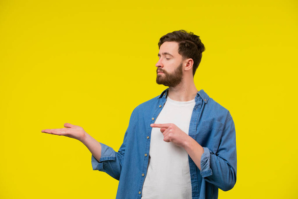 Portrait of a young adult man in blue shirt holding his arm up with open palm. Pointing his finger at the hand and looking at it. Space for inserting object. Isolated on yellow background. - Photo, image