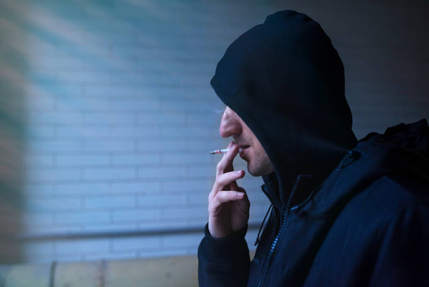 Close-up portrait of a man with a cigarette and a hood. Social issues, drug addiction and crime. - Fotoğraf, Görsel