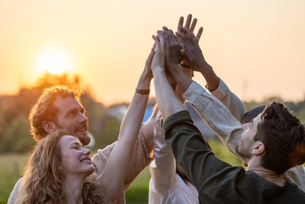 Multiracial group of diverse young people giving high five against a setting sun, feels excited close up focus on stacked palms during golden hour. Respect and trust, celebration and friendship - Photo, image