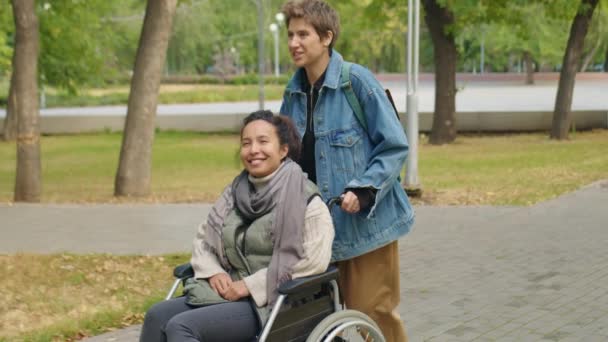 Mittlere Langzeitaufnahme einer fröhlichen jungen Frau, die eine Freundin im Rollstuhl schubst und mit ihr bei einem Spaziergang im Park an einem Herbsttag plaudert - Filmmaterial, Video