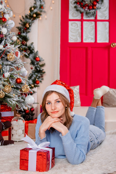 A young, attractive woman lies with a gift in her hands against the background of a Christmas tree and New Year's decorations. - Foto, imagen