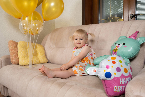 Kaksivuotias blondi tyttö juhlii syntymäpäiväänsä istuu sohvalla ilmapallojen joukossa.. - Valokuva, kuva