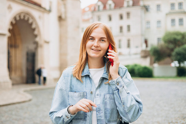 Schöne lächelnde Frau, die am Telefon spricht und draußen steht. Telefonische Kommunikation. Glückliche, fröhliche junge Frau, die auf der Stadtstraße läuft, urbanes Lifestyle-Konzept. - Foto, Bild