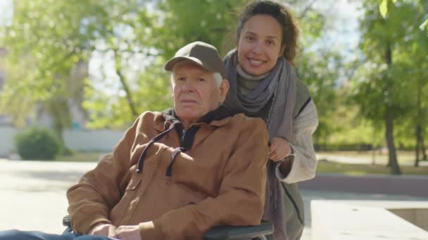 Portret uśmiechniętej opiekunki i starszego mężczyzny na wózku inwalidzkim pozujących razem do kamery na spacerze w parku - Materiał filmowy, wideo