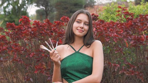 jonge vrouw in een groene jurk houden make-up borstels tegen een achtergrond van bloemen - Foto, afbeelding