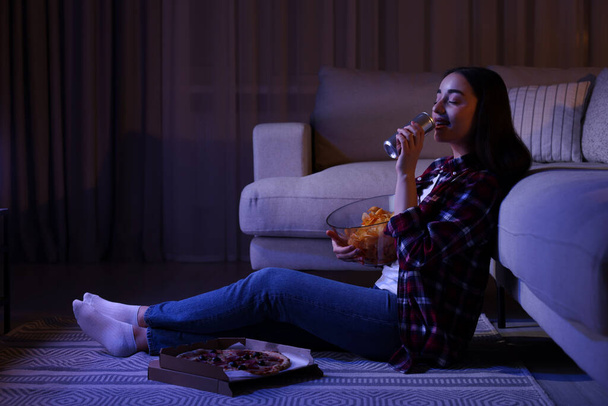 Young woman drinking from tin can and eating chips while watching TV in room at night. Bad habit - Zdjęcie, obraz
