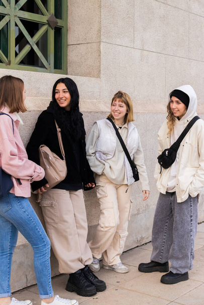 Quatre étudiants amis, multiraciaux, jeunes femmes, trois caucasiens et une brune, bavardant à l'extérieur de l'université, vêtus de vêtements d'hiver, avec un mur blanc en arrière-plan. - Photo, image