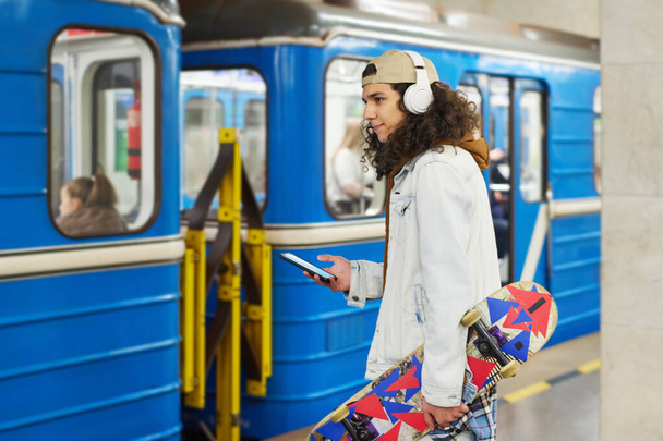 Boční pohled na moderní teenager ve sluchátkách stojící před modrým metrem vlaku a pomocí mobilního telefonu, zatímco se chystá cestovat metrem - Fotografie, Obrázek
