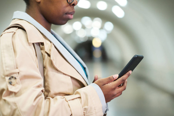 Cropped shot of side view of black woman in beige trenchcoat holding smartphone while texting in front of camera in subway tunnel - Foto, Imagen