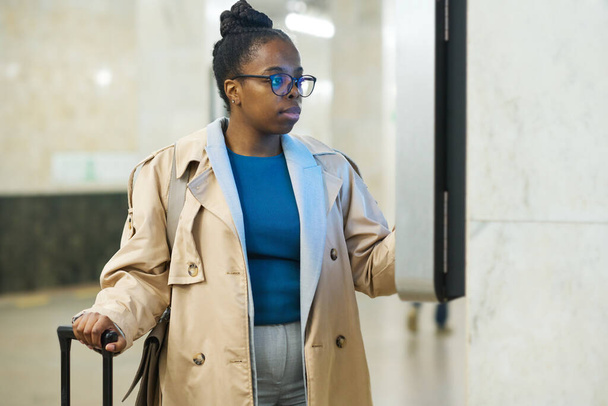 Молода серйозна жінка-мандрівник в окулярах і бежевий тренч, стоячи перед дисплеєм і переглядаючи розклад поїздів метро - Фото, зображення