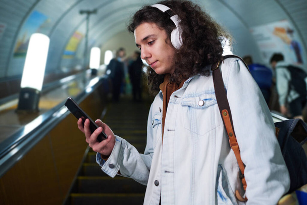 Teenage guy v casualwear a sluchátka pomocí smartphone zatímco stojí na eskalátoru, sledování videa a pohybující se dolů - Fotografie, Obrázek