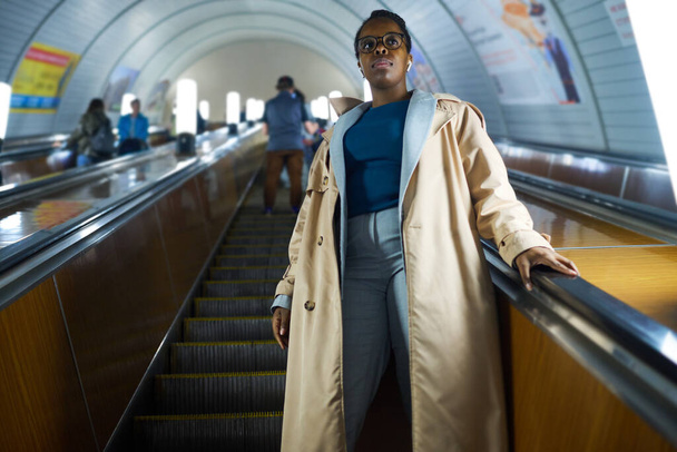 Молода афроамериканка в повсякденному одязі стоїть на ескалаторі і рухається вниз проти довгого тунелю метро з іншими людьми - Фото, зображення
