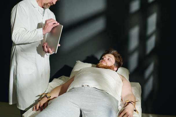 Ώριμος γιατρός με εργαστηριακή ποδιά που δείχνει κάτι στην οθόνη του δισκίου σε νεαρό άνδρα ασθενή δεμένο στο κρεβάτι με σφιχτές ζώνες κατά τη διάρκεια της κλινικής θεραπείας - Φωτογραφία, εικόνα