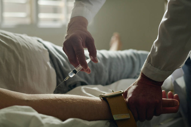 Close-up of doctor of lunatic asylum making injection to young male patient with seasonal affective disorder lying in bed in hospital ward - Valokuva, kuva