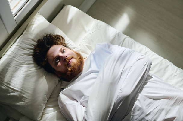 Над углом юного бородатого мужчины в смирительной рубашке, страдающего шизофренией, лежащего на кровати под психиатрической больницей - Фото, изображение