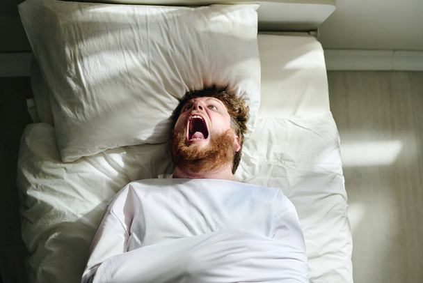 Young insane man with psychosis in straitjacket shouting while lying on bed in mental hospital during therapy recommended by his doctor - Foto, Bild