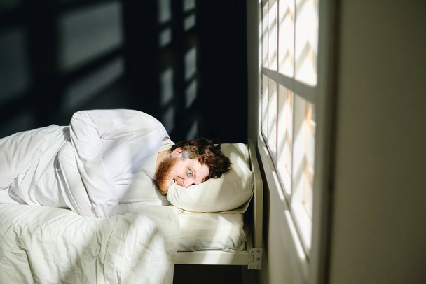 Joven hombre trastornado con camisa de fuerza mordiendo almohada mientras está acostado en la cama por ventana en el hospital psiquiátrico en aislamiento durante la terapia - Foto, imagen