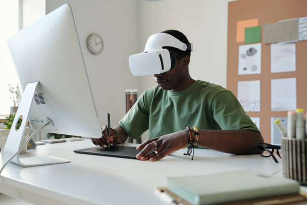 Joven diseñador masculino en realidad virtual auriculares dibujo bocetos gráficos en el panel táctil con lápiz mientras está sentado frente a la pantalla de la computadora - Foto, Imagen