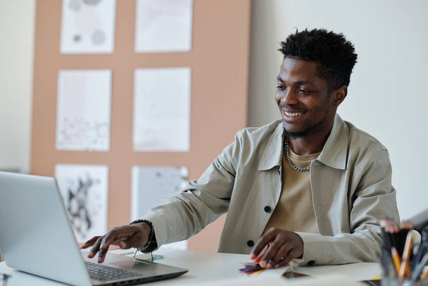 Giovane sorridente webdesigner afroamericano maschio premendo il tasto della tastiera del computer portatile mentre seduto sul posto di lavoro e la creazione di nuovo sito web - Foto, immagini