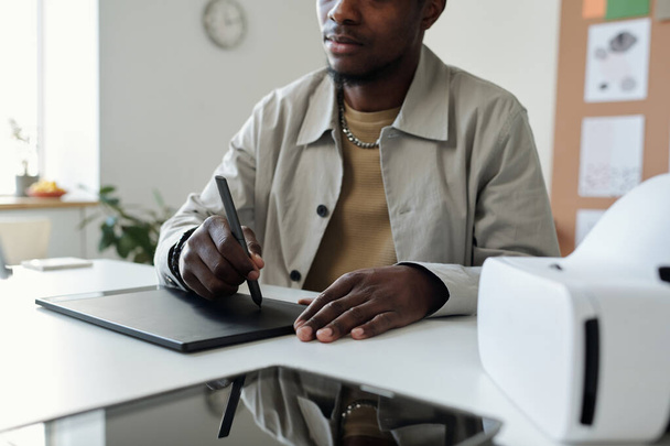Oříznutý snímek mladé afroamerické mužské designérky kreslící grafiku při sezení na pracovišti a držení stylus na dotykovém displeji - Fotografie, Obrázek