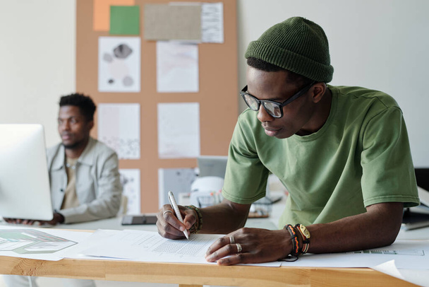 Νεαρός Αφρο-Αμερικανός σχεδιαστής ή αρχιτέκτονας σε casualwear και γυαλιά σχεδίασης σκίτσο του νέου κτιρίου στο γραφείο, ενώ κάμψη πάνω από χαρτιά - Φωτογραφία, εικόνα