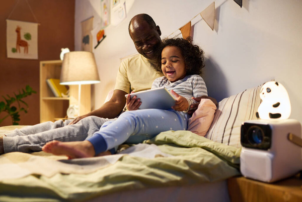 Afroamerikanisches Kind, das zusammen mit seinem Vater vor dem Schlafen in der Nacht lustige Zeichentrickfilme auf dem digitalen Tablet anschaut - Foto, Bild