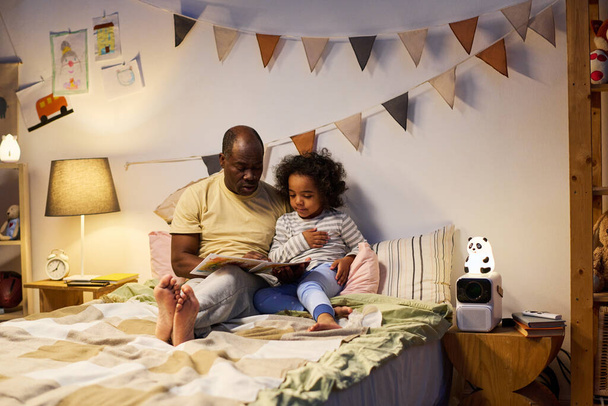Afroamerikanischer Vater liest seinem Sohn vor dem Schlafen Buch vor, während sie im Schlafzimmer auf dem Bett sitzen - Foto, Bild