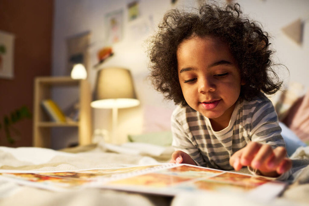 Afro-Amerikalı çocuk yatakta yatarken uyumadan önce kitaptan resimler izliyor. - Fotoğraf, Görsel