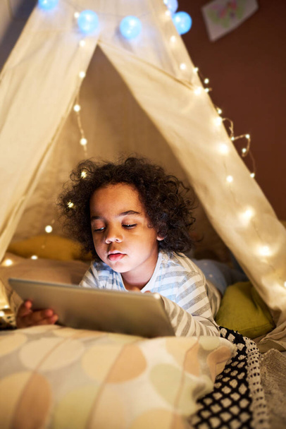 Little boy using tablet pc to watch cartoon while lying in handmade tent at home - Fotografie, Obrázek