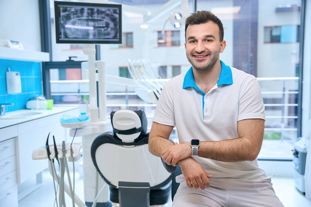 Усміхнений чоловік сидить на робочому місці в стоматологічному кабінеті, лікар має розумний годинник - Фото, зображення