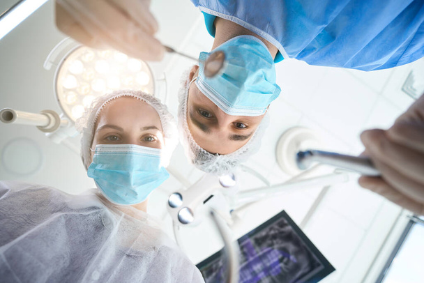 Chirurgo con uno specchio dentale tra le mani sul posto di lavoro in sala operatoria, un assistente femminile lo aiuta - Foto, immagini