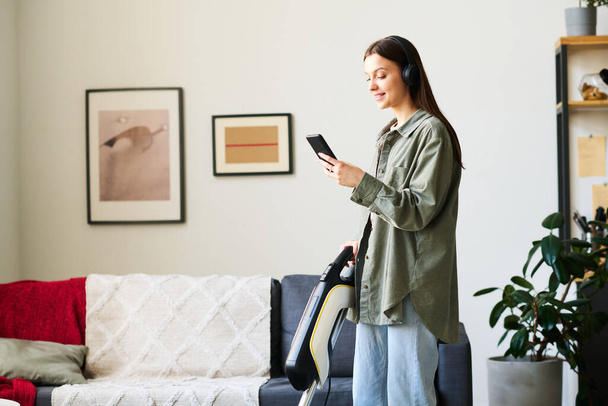 Young woman in headphones using smartphone to listen to music while vacuuming at home - Foto, imagen
