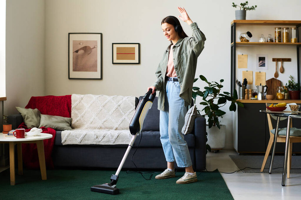 Woman in headphones listening to music and dancing while vacuuming the carpet in the room - Foto, afbeelding