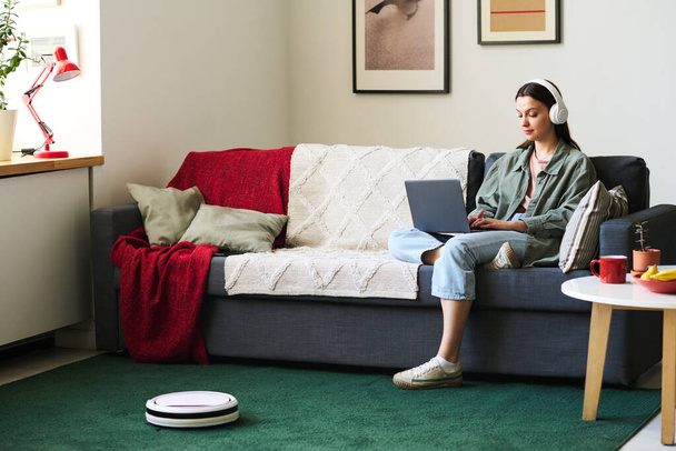 Jovem mulher trabalhando on-line no laptop sentado no sofá durante o trabalho doméstico com aspirador de pó robô - Foto, Imagem