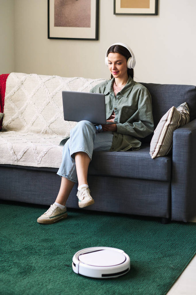 Fiatal nő fülhallgatóban laptoppal ül a kanapén, miközben robot porszívó tisztítja a szőnyeget - Fotó, kép