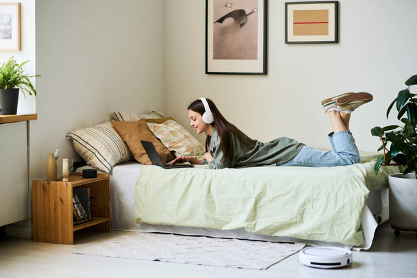 Young woman in headphones using laptop while lying on bed in her bedroom - Photo, Image