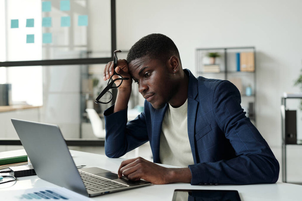 Young tense African American businessman sitting by workplace in front of laptop in office and looking at screen with data while analyzing it - 写真・画像