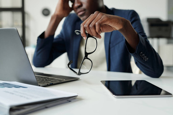 Focus on hand of young pensive African American businessman holding eyeglasses while sitting by desk with laptop and tablet in office - Foto, imagen