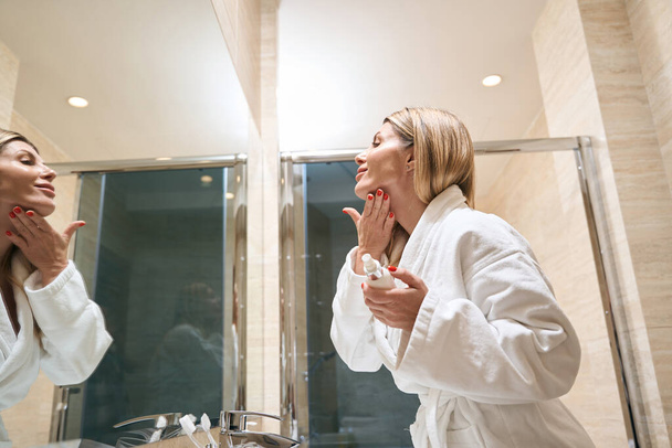 Female in white coat standing near mirror and holding bottle, touching skin in the hotel - 写真・画像