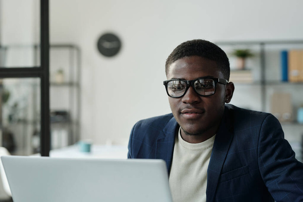Giovane serio collare bianco in occhiali seduto di fronte al computer portatile e guardando lo schermo durante la comunicazione online in video chat - Foto, immagini